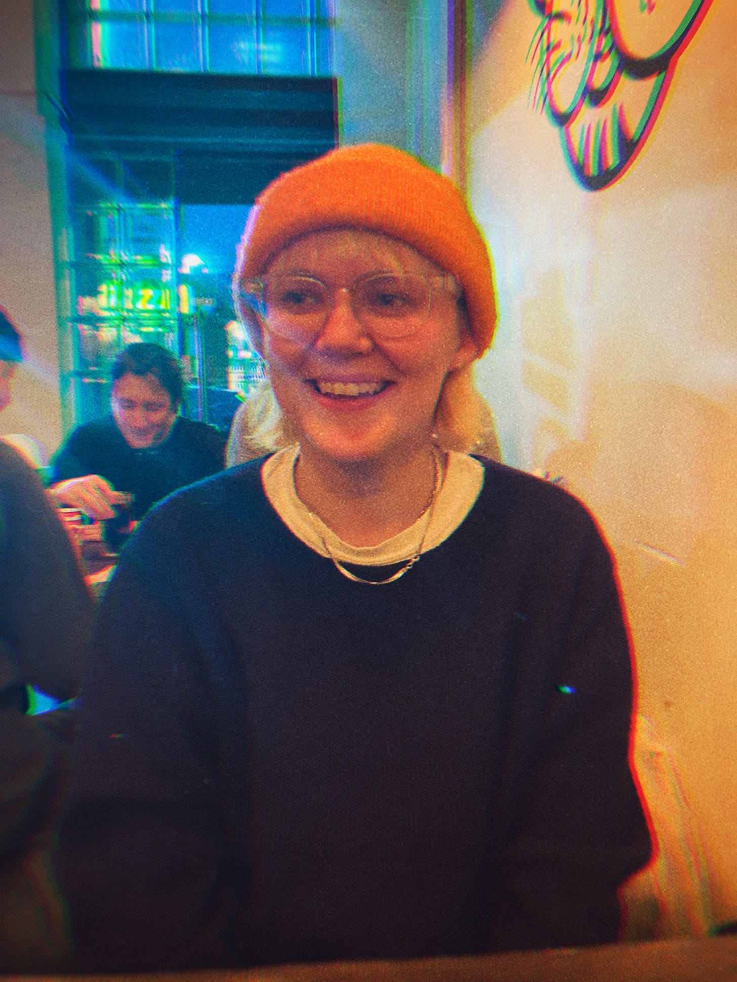 A portrait photo of a person sat opposite a table in restaurant, they wear a navy jumper with a white t-shirt underneath and gold necklace on top, they also wear an orange beanie that covers blonde chin length hair and they wear large round shaped see-through glasses. They are smiling and looking off to the left.