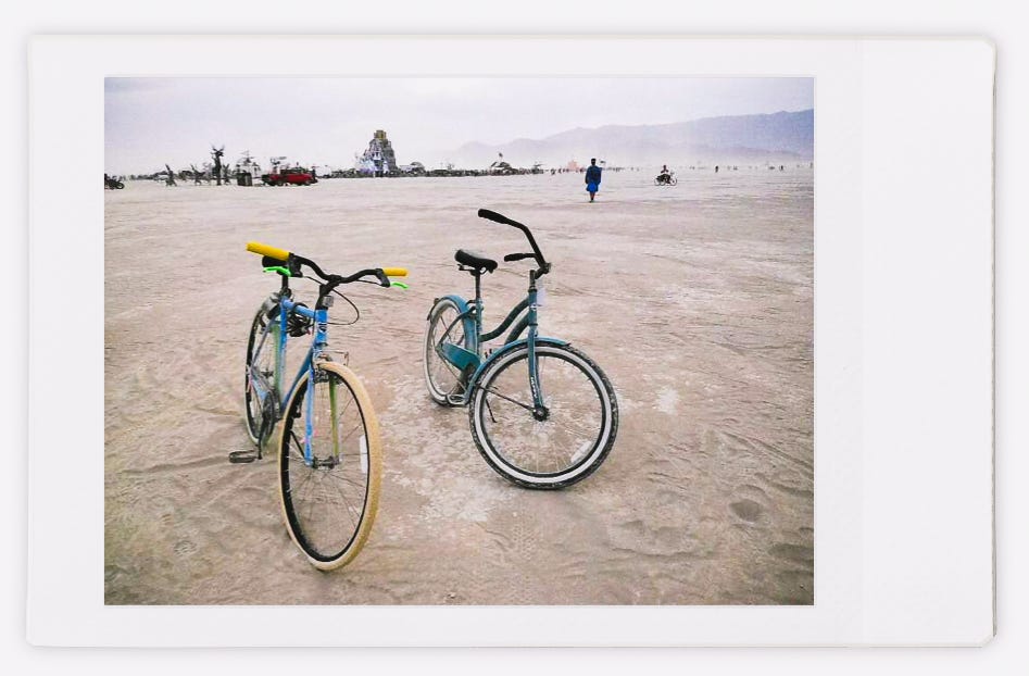 Two bikes parked in the desert, various art pieces in the middle ground, and the mountains behind them
