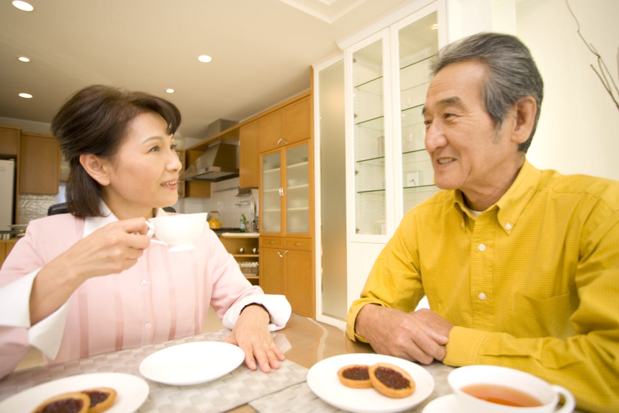 Middle-aged Asian couple sitting at kitchen table in new living quarters. Wife holding cup of coffee. Four tarts on table.