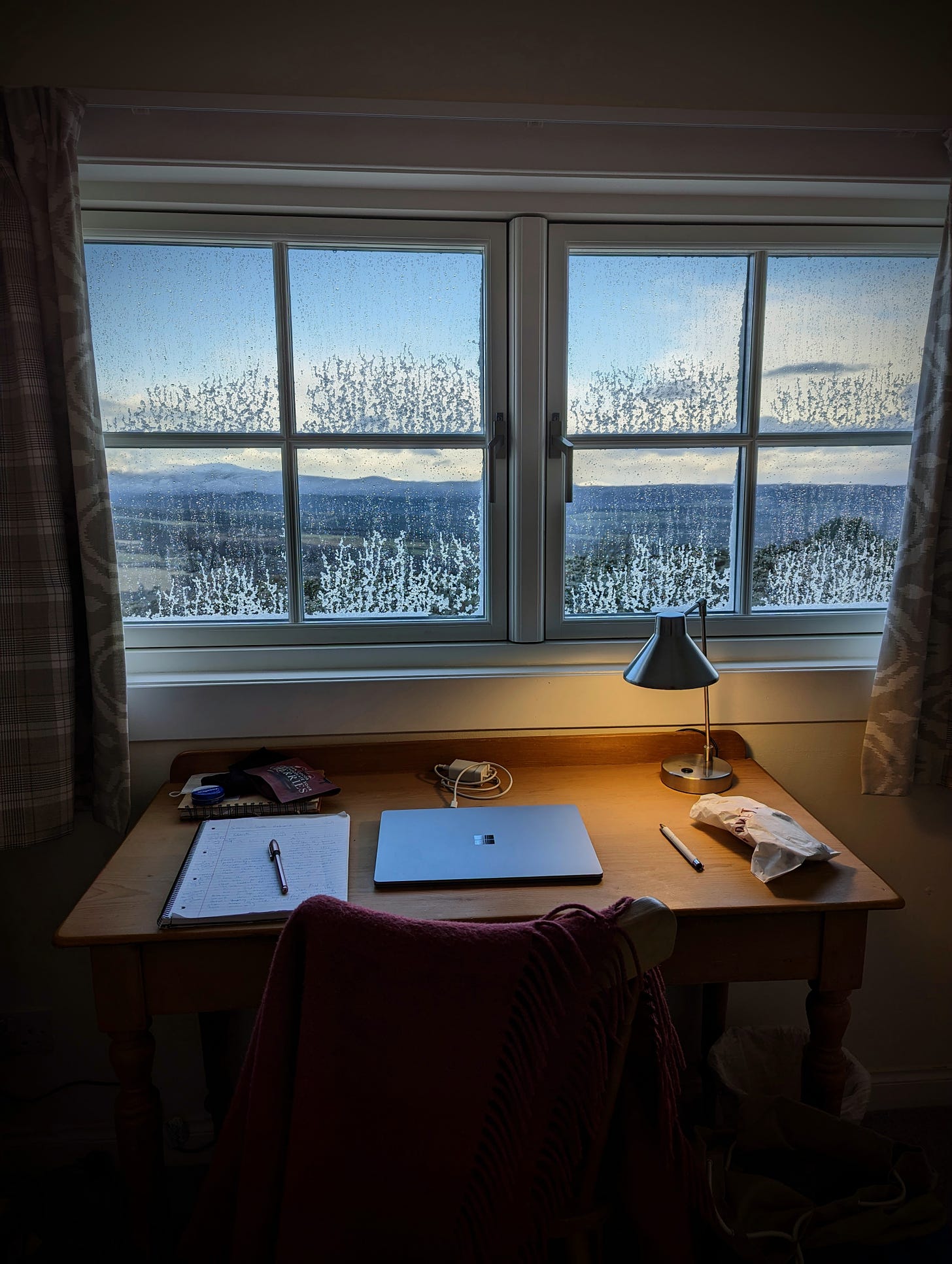 A photo of a wooden desk under a cottage window. The desk has a laptop, a lamp casting a warm glow, a notepad and pen, and some oddments. A chair has a cosy wool blanket thrown over it. Through the window, a valley and distant hills can be seen, and the glass is patterned with frost. 