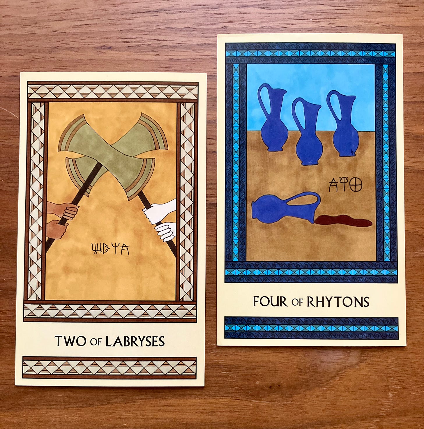 Two Minoan Tarot cards side by side on a wooden table. The Two of Labryses is in shades of gold and cream. It shows two pairs of hands holding large, equally-sized labryses that are clashing. The Four of Rhytons is in shades of blue and tan. It shows three pitchers sitting on a table with a fourth pitcher tipped over, its contents spilling out.