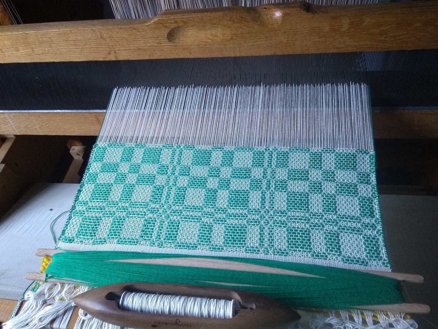 The very start of hand-woven fabric on a floor loom, with a geometric checkered pattern in green and white. A stick shuttle and a boat shuttle are resting at the bottom.