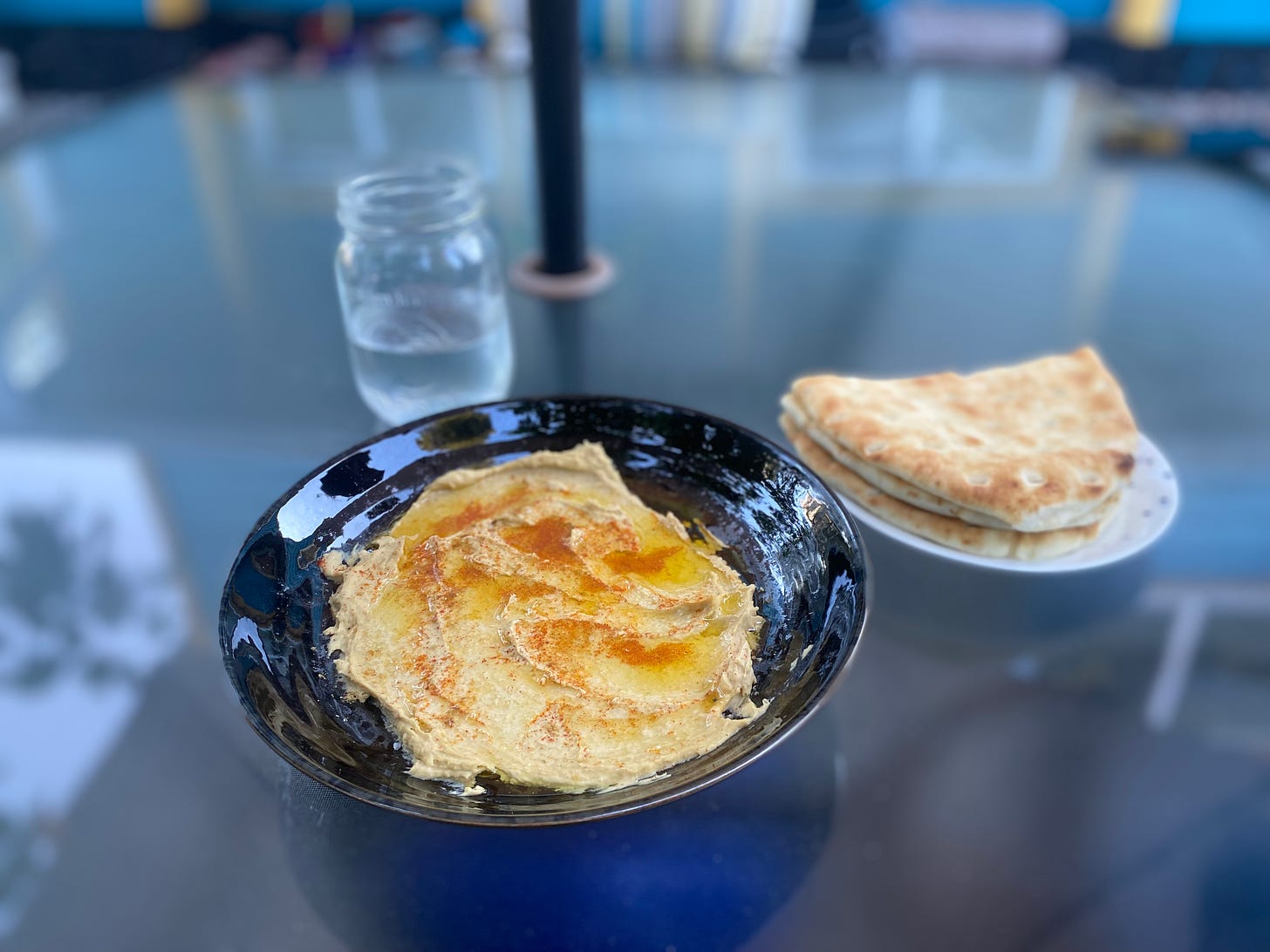 A shallow black bowl filled with a spread of hummus, topped with olive oil, smoked paprika, and cumin. On a small plate are slices of naan, and in the background is a glass of water in a mason jar.