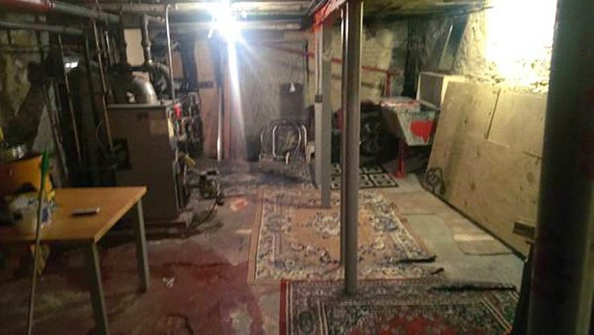 A scary, dark basement apartment lit by a glaring bare lightbulb. lots of pipes going to and from a furnace on one side of the room. on the other, a laundry sink has a lopsided cabinet balanced above it, with a mirror sitting atop the cabinet.  The unfinished cement floor has a couple of very old, worn carpets on it. Very serial-killer vibe.