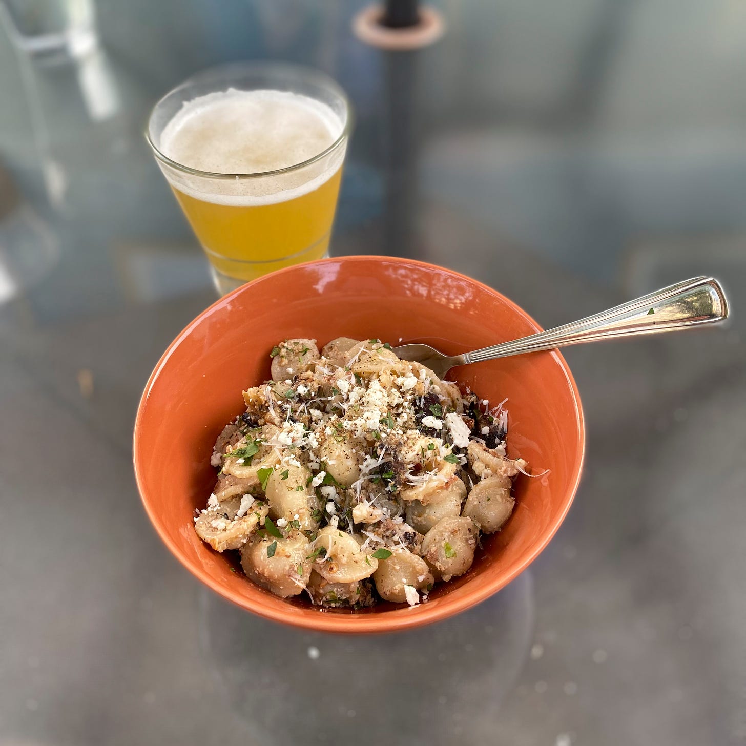 On an outdoor glass table, an orange bowl of the pasta salad described above, dusted with feta, parmesan, herbs, and black pepper. A fork sticks out of the bowl on the right, and a glass of hazy beer is visible in the background.