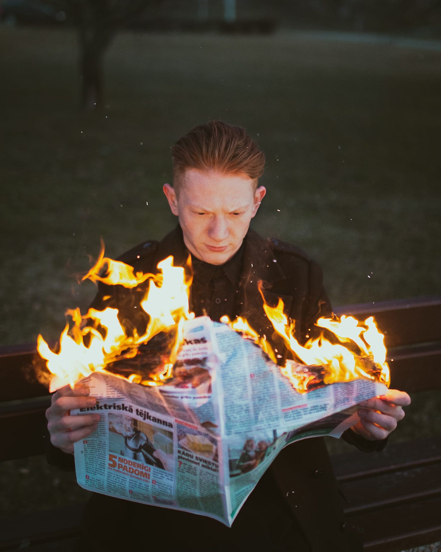 A picture of a red-haired white man sitting on a bench in Riga, Latvia and trying to read a Latvian newspaper that is literally on fire and burning up in his hands.