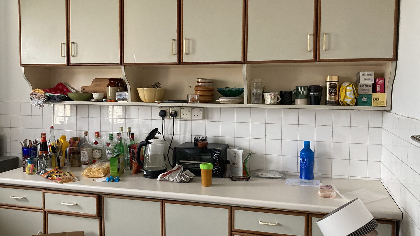 A set of large built-in kitchen cabinets. On an open shelf, plates, bowls, clothes and mugs are artfully arranged. On the countertop, there is a mess of things: jars, glass gin bottles, a clear kettle, a toaster oven, knives and forks in a waffle tin, and all manner of plastic-wrapped packages.
