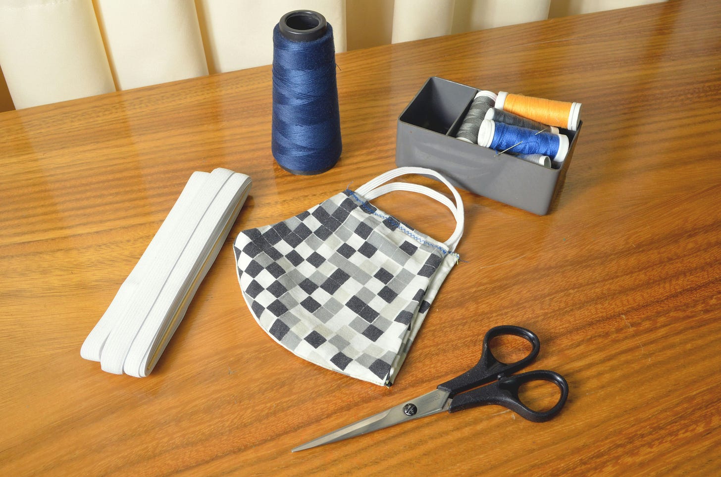 a wooden table with sewing and crafting supplies on top - scissors, clothing elastic, a spool of blue thread, a bin with other colors of thread, a small grey checkered bag with a white handle