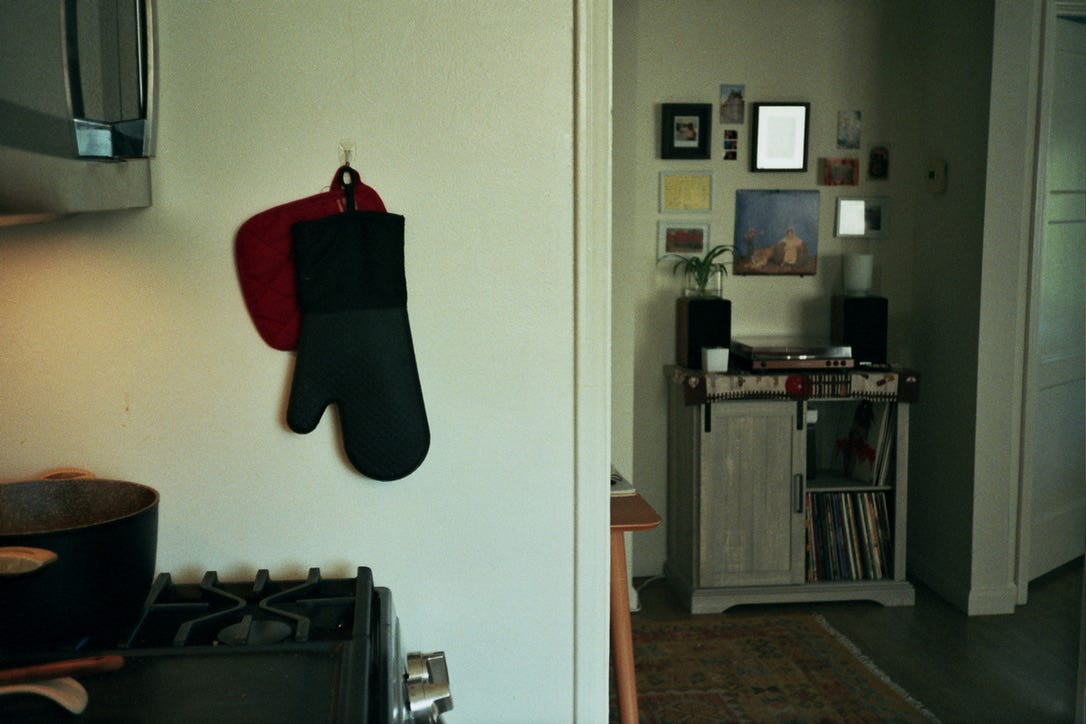 In the left side of the picture, foregrounded is a gas stove and microwave. There's a pot on the stove, a spoon holder, and two oven mitts hanging from a hook on the wall. The right side of the picture shows a cabinet with a sliding door and lots of vinyl records stacked vertically. A record player and speakers sit on top. There are many small images and frames on the wall above it, and a colorful rug below.