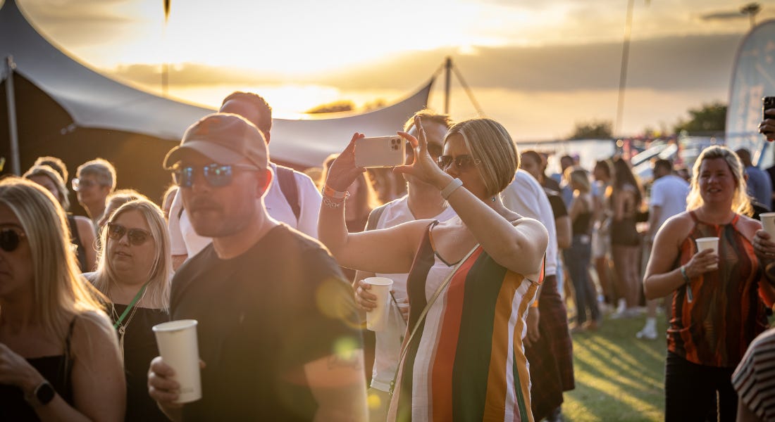 A crowd of people at a festival, including a woman taking a picture of her phone, with a sunset in the background
