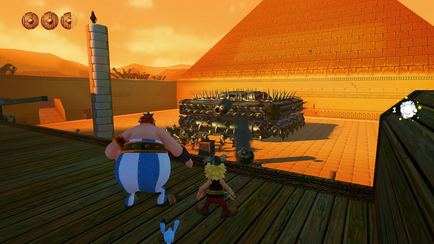 Screenshot from the game. Asterix and Obelix look down on a Roman war machine covered in spikes, with a troop of soldier in front of it. A pyramid looms in the background.