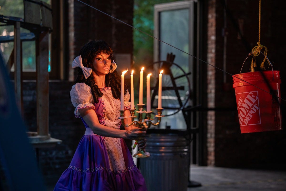 Narcissister stands in a dim warehouse, holding a candelabra that illuminates her face. She has light brown skin and wears a mannequin mask, a white and purple dress, and bows in her hair.