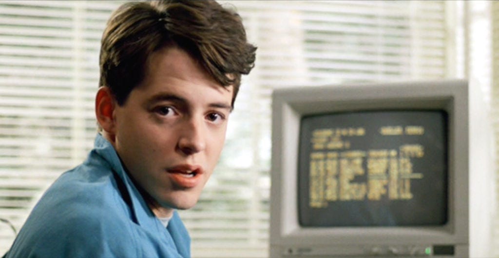 Ferris Bueller breaks the fourth wall, looking at the camera while seated in front of a computer monitor in his bedroom.