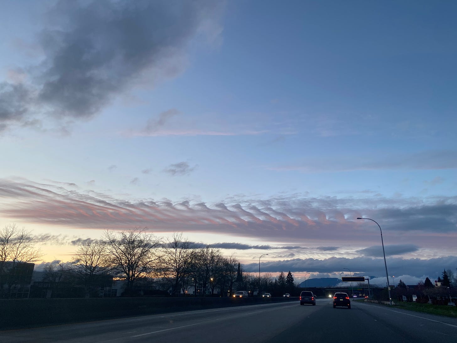 A view of the sky from the highway after Sunday's storm. It's near sunset, and the sky is a deep blue, with rippling clouds tinged pink and orange near the horizon. Cars on the highway have their lights on, and the mountains are just visible in the distance.