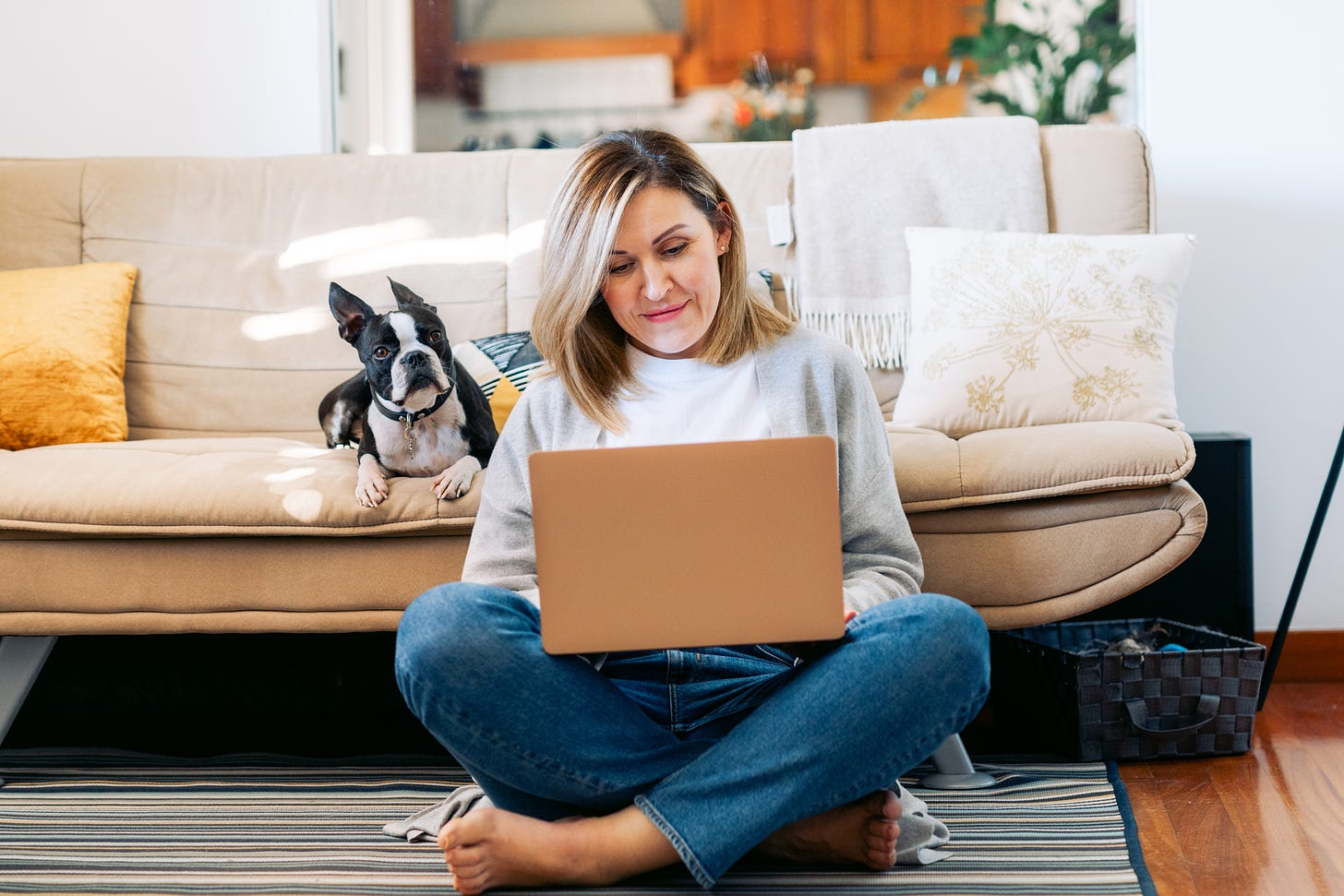 A woman works on her laptop while sitting next to her dog in her living room.