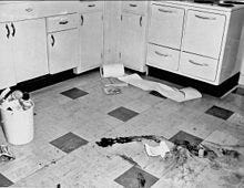 A black and white image of a checkered tiled floor with a right-angled row of light-colored cabinets at the rear. On the floor in the foreground are several large dark smears, with a small piece of paper on the ground nearby. At the left is a small white plastic bucket filled to over the top with trash; some cans and a telephone handset are visible. In the rear, near the cabinets, is a roll of paper with part of it spilling off to the right; underneath it are what appear to be two books