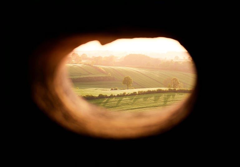 A hole in a wall that shows an idyllic green field and ‘greener pastures’ on the other side