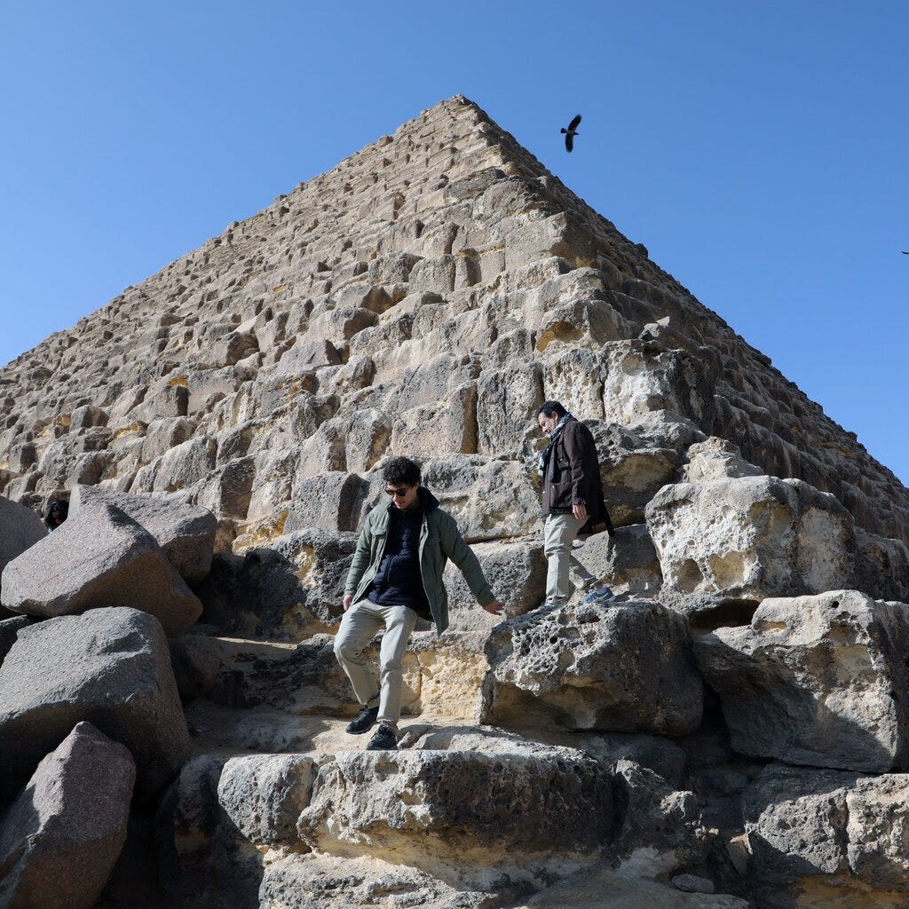 Two men climbing on a pyramid in front of a blue sky. 