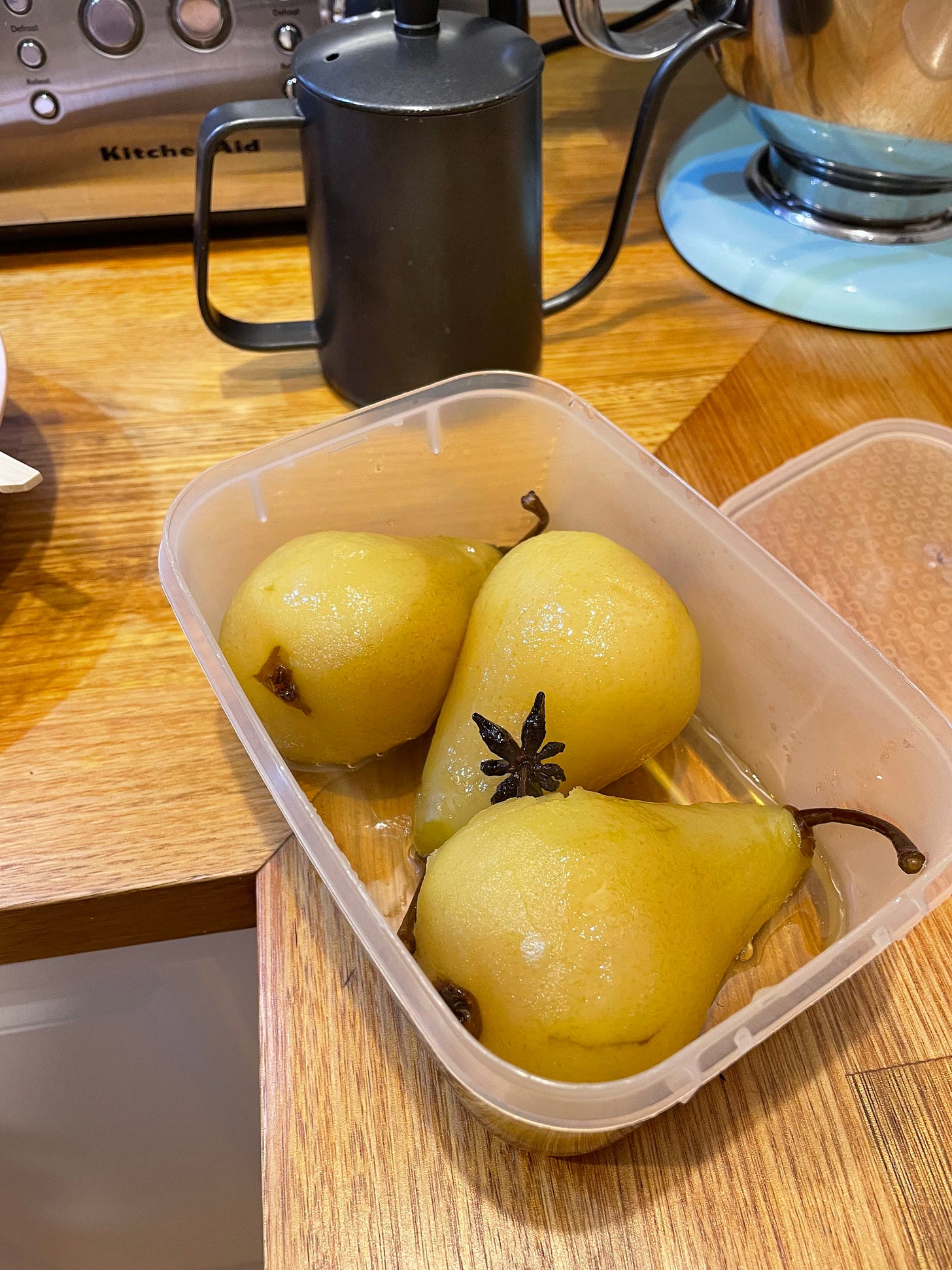 Three poached pears lying down in a container with syrup ready to go in the fridge. A gooseneck kettle in the background.