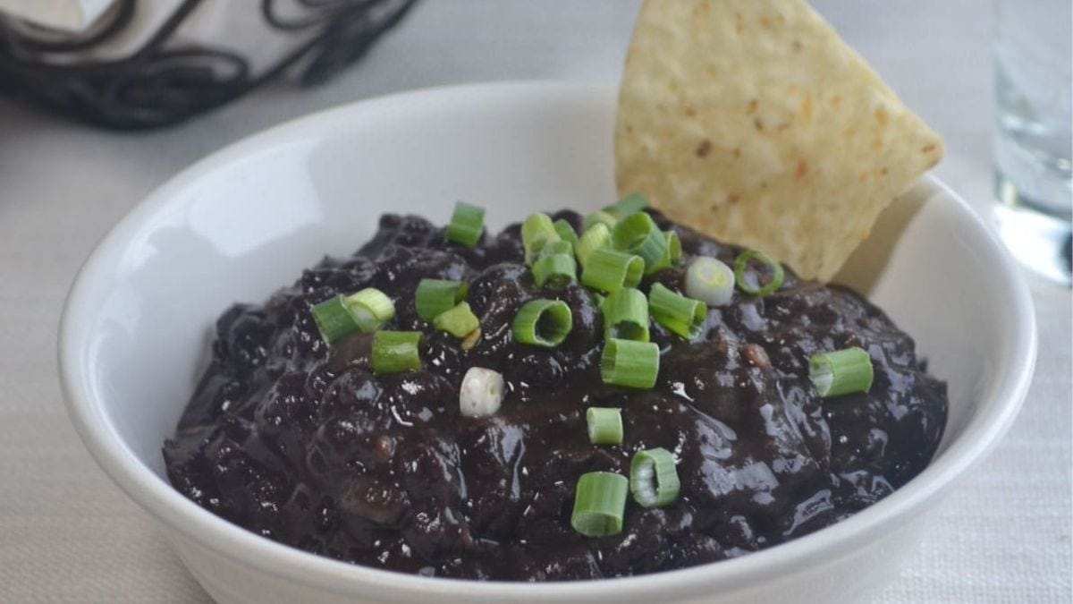 A bowl of black bean soup garnished with chopped green onions, accompanied by a piece of tortilla chip on the side, set on a light-colored tablecloth.
