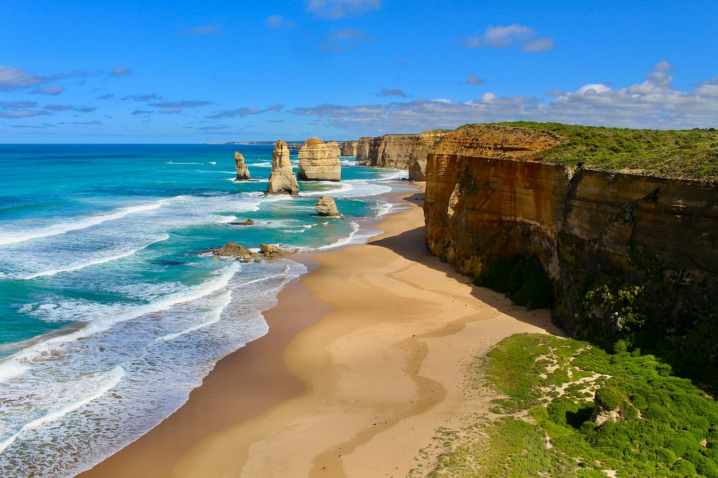 Beach scene. The stark cliffs off the Great Ocean Road give way to pristine sandy beachs and blue ocean. In the background several sea stacks can be seen coming out of the water. Beach scene. The stark cliffs off the Great Ocean Road give way to pristine sandy beachs and blue ocean. In the background several sea stacks can be seen coming out of the water.