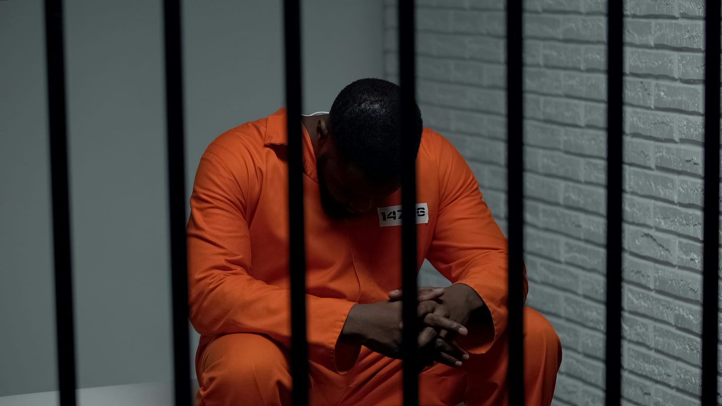 Black incarcerated man in orange jumpsuit with head down behind prison bars