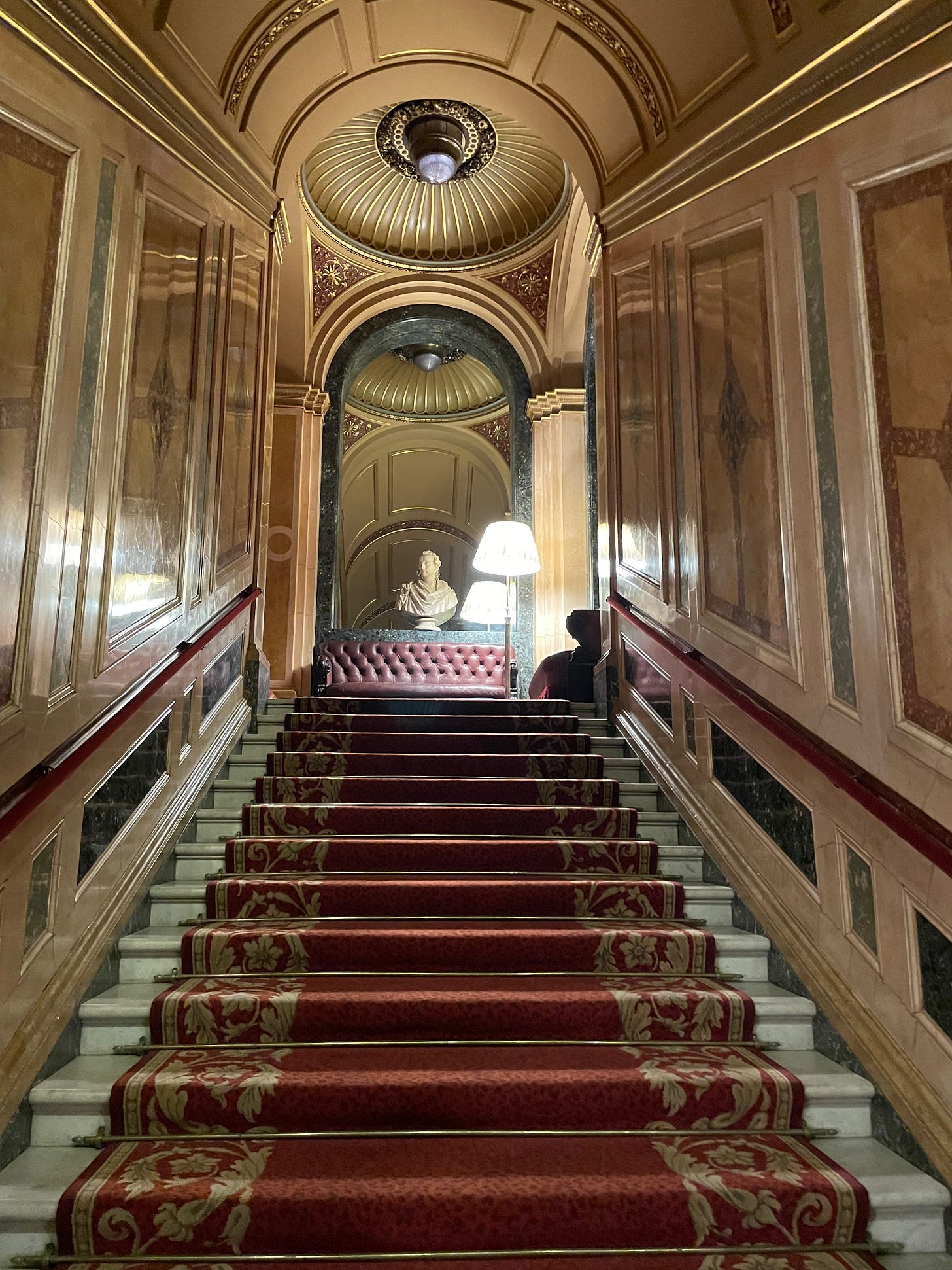 A grand staircase: red carpet, gold walls, and a marble bust at the top.