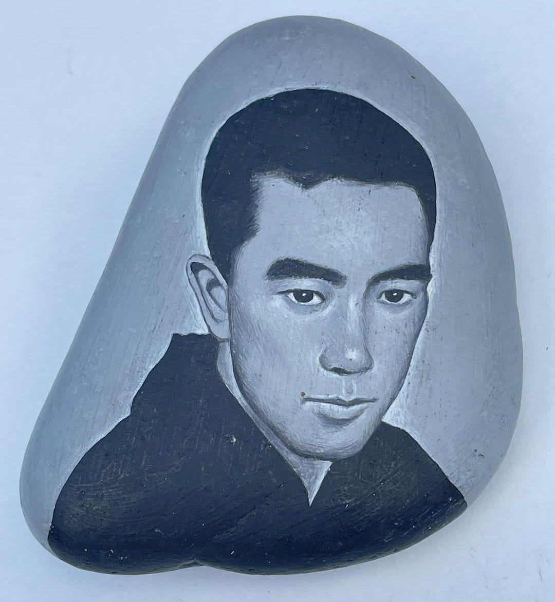 A light blue background with an oblong stone in the center is pictured. There is a black and white painting of Yukio Mishima in the middle of the stone. He has short black hair and a black shirt. A light blue background with an oblong stone in the center is pictured. There is a black and white painting of Yukio Mishima in the middle of the stone. He has short black hair and a black shirt.