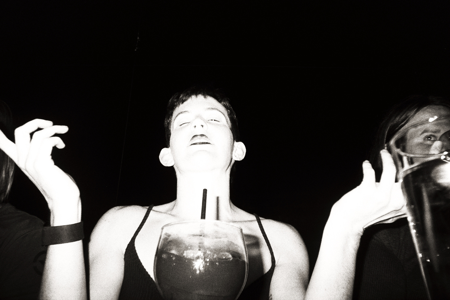 Black and white photograph of Lyss with their eyes closed and hands outstretched, wearing black lipstick and a black top. The photo is grainy and has a vintage vibe to it. The background is completely black, and the camera flash has illuminated the foreground to complete white.