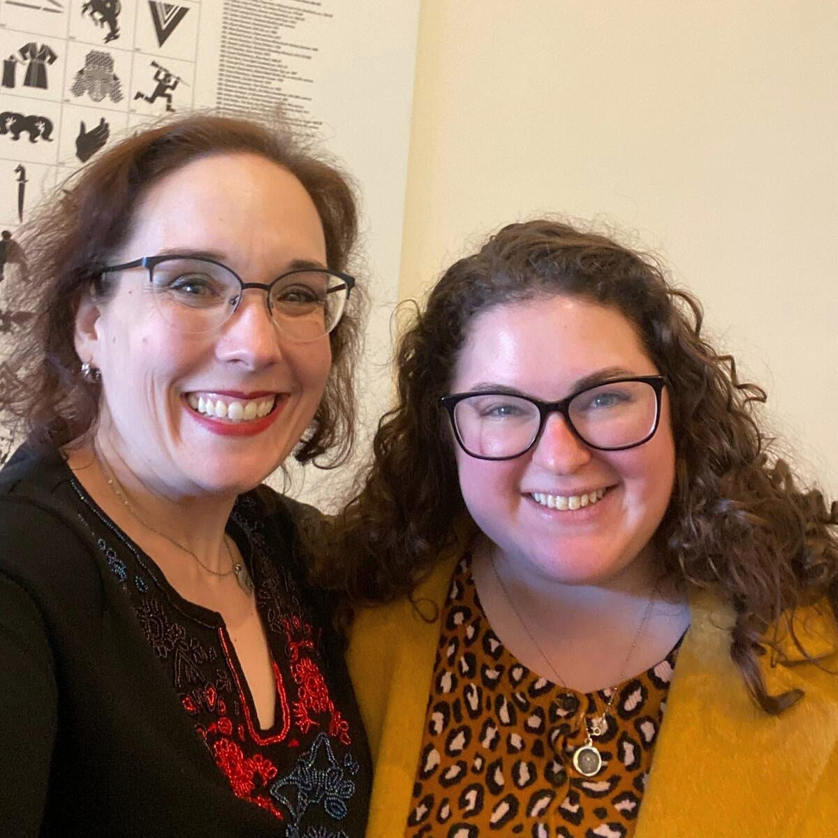 Audrey (a white woman in her 40s with dark red short hair and glasses) and Sarah (a white woman in her 30s with curly brown hair and glasses) stand close together smiling at the camera.