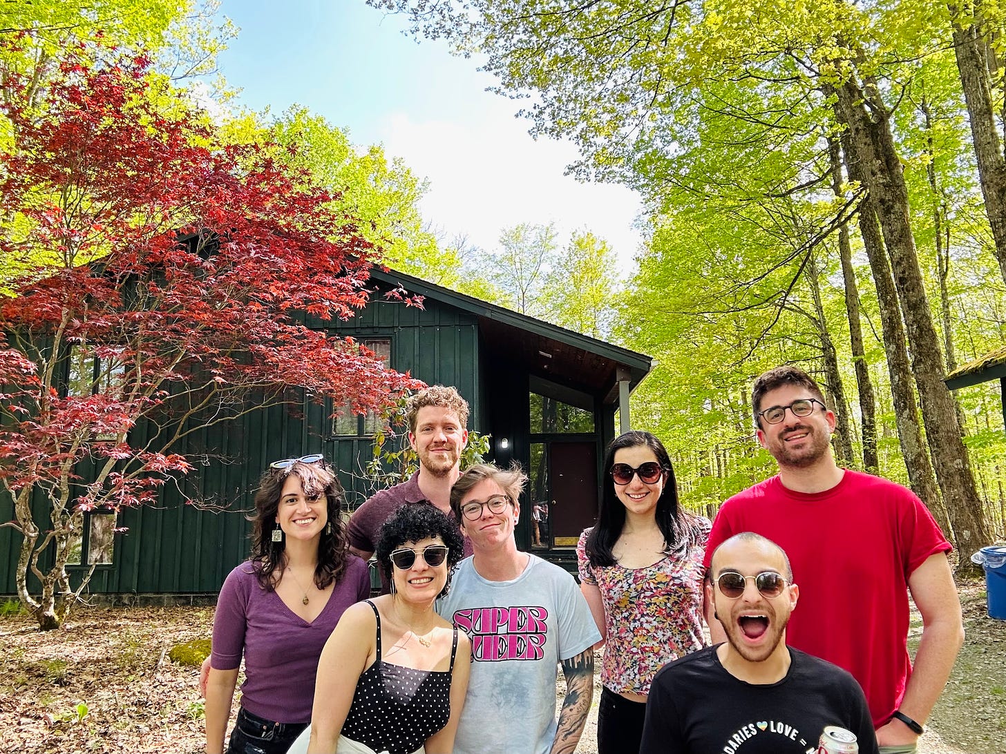 A group of people smile at the camera standing in two lines in front of a blue house surrounded by trees