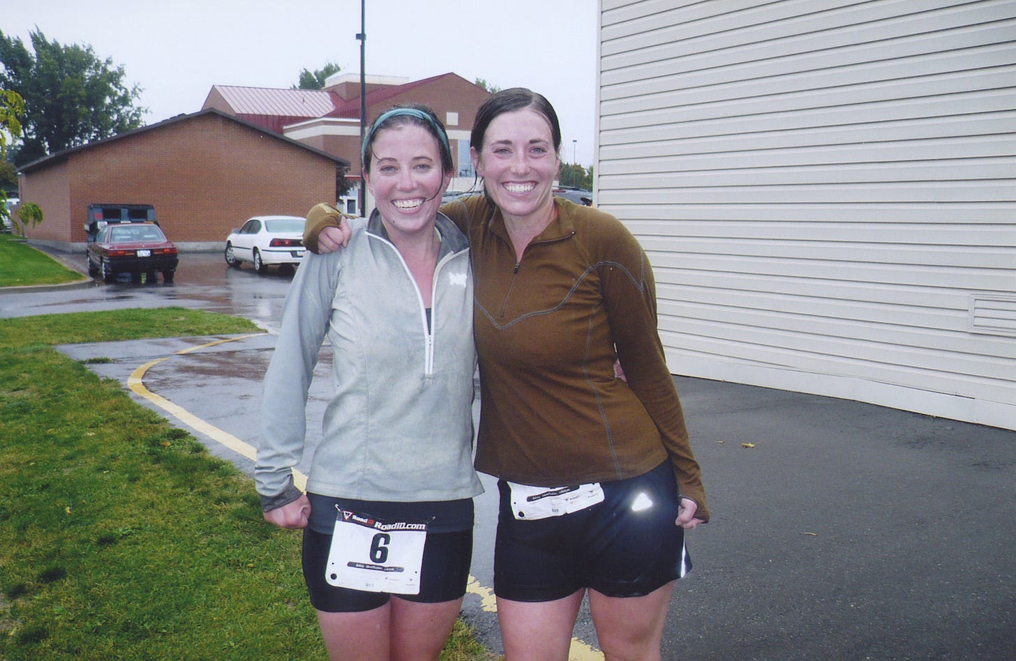 Two young women, wet from the rain, in  running jackets, shorts, and race bibs, smile, arm in arm.