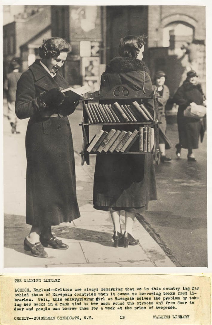 A black and white photo from the 1930s of two women standing in a street. One has a small bookshelf strapped to her back and the other is holding a booking and reading it. There’s a caption beneath the image that reads: Critics are always remarking that we in this country lag far behind those of European countries when it comes to borrowing books from libraries. Well, this enterprising girl at Ramsgate solves the problem by taking her books in a rack tied to her back round the streets and from door to door and people can borrow them for a week at the price of twopence.