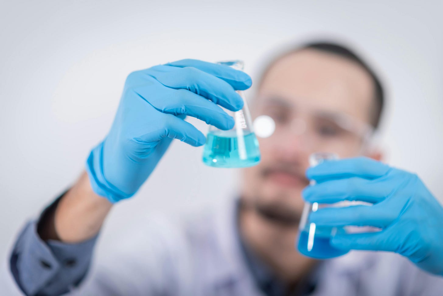 Photo of a man wearing safety goggles and a lab coat, whose face is blurred in the background. The foreground focuses on his hands (wearing blue latex gloves) holding two small flasks containing slightly different shades of blue liquid. It is obviously a staged photo rather than depicting actual science.
