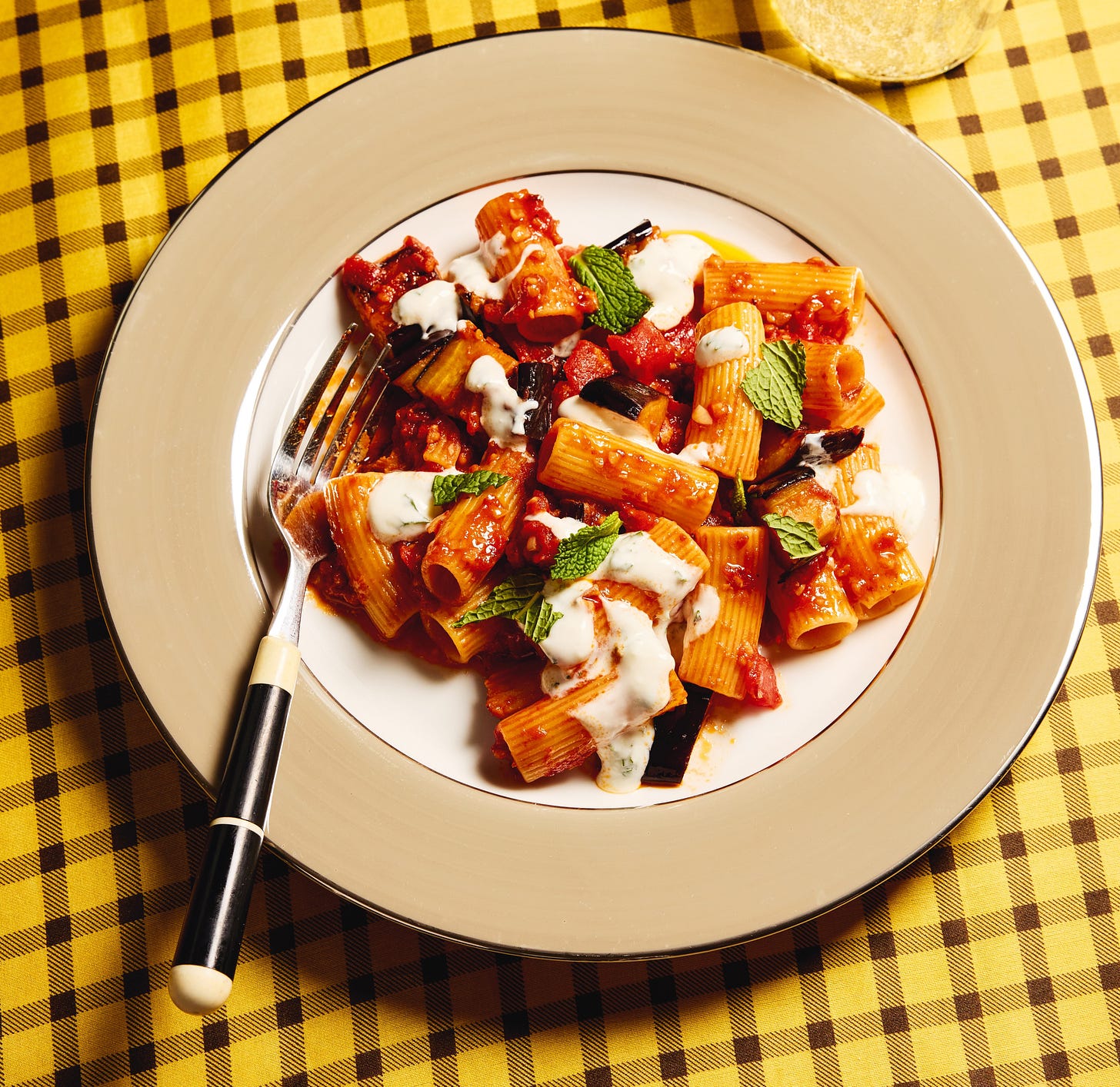 Plate of tomato sauced pasta with eggplant and yogurt on yellow checked tablecloth.