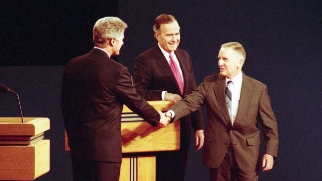 Bill Clinton shakes hands with Ross Perot in front of a lectern while George H. W. Bush looks on.