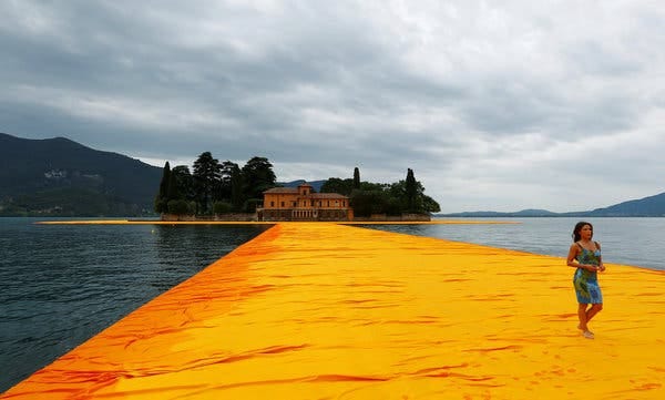 A new installation by the artist Christo, “The Floating Piers,” consists of temporary bridges spanning Italy’s Lake Iseo. A woman in a blue dress walks across the golden fabric on the water in this image, the sky grey behind her.