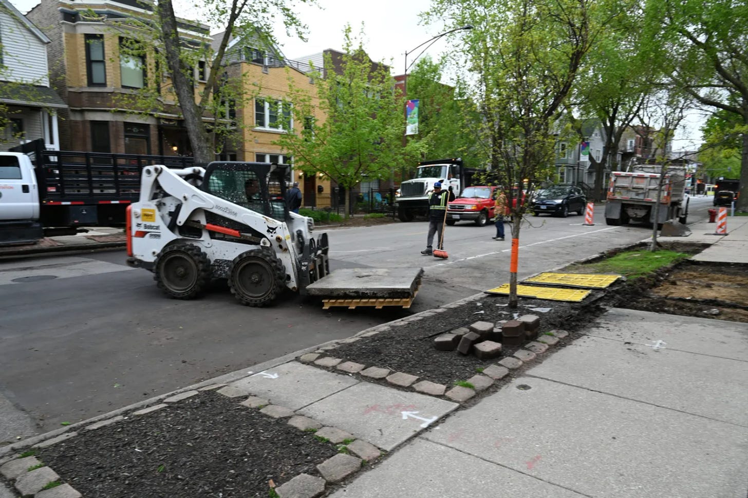 A city street in Chicago. A bobcat has the entire concrete slab containing the rat hole on the front and is wheeling it away from its original location. That area is now a dirt patch in the sidewalk.