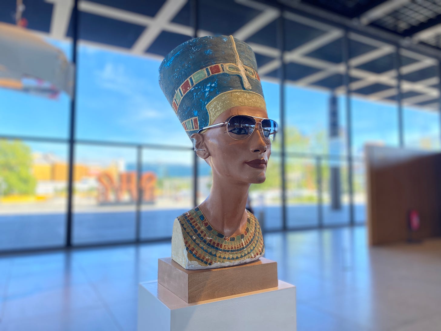 Imagem: Cópia busto Nerfetiti usando óculos aviador exposto numa sala ampla com uma parede de vidro aos fundos e céu azul.