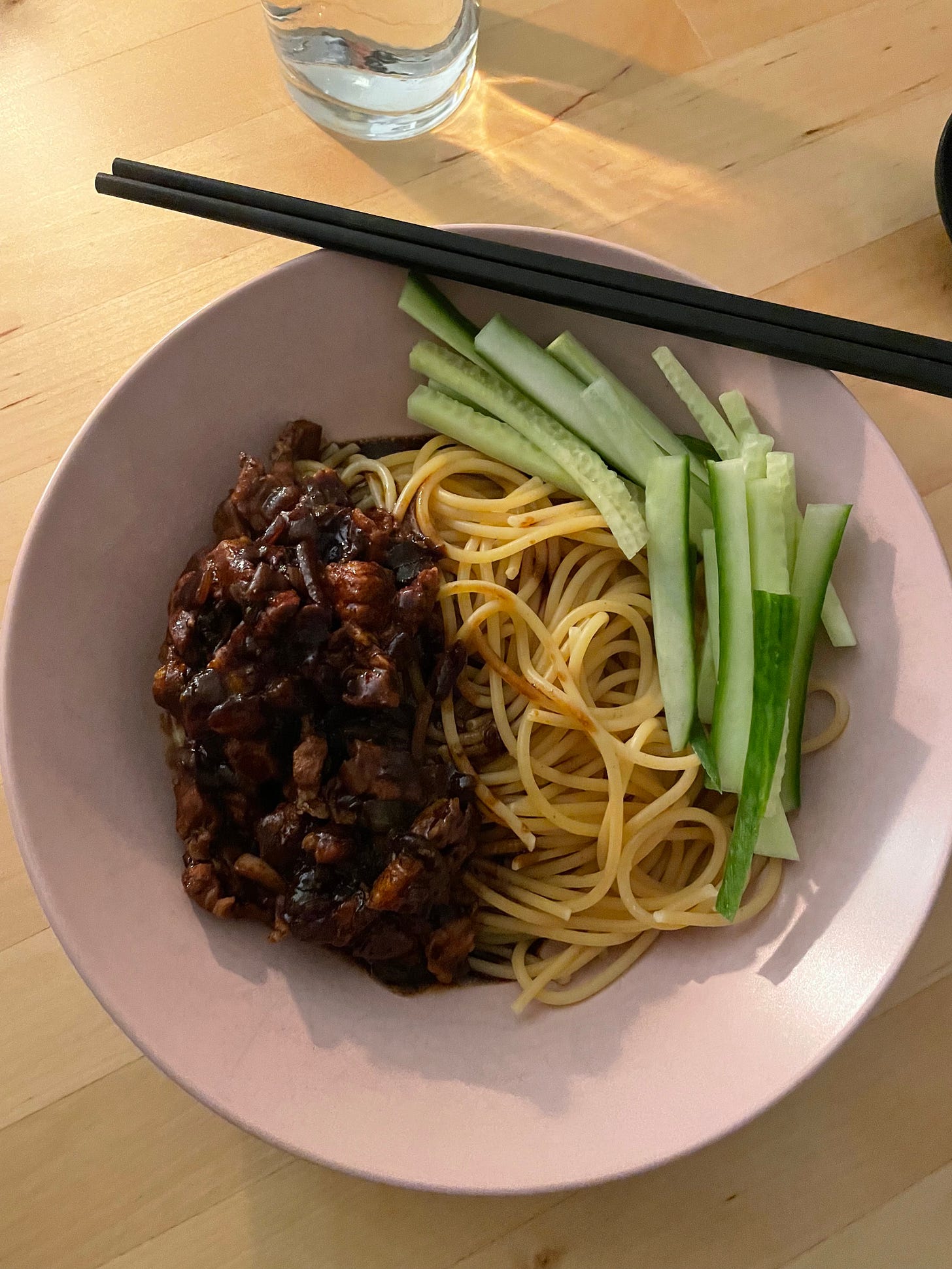Pink bowl filled with spaghetti and a black bean sauce with pork belly. Sliced cucumber on the side, a Korean dish called Jajanmyeon.