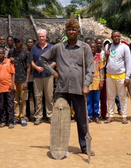 The chief of Baringa village in a long-sleeved shirt, holds a spear and has a shield propped up on his right leg outside his hut with a crowd of people behind him The chief of Baringa village in a long-sleeved shirt, holds a spear and has a shield propped up on his right leg outside his hut with a crowd of people behind him