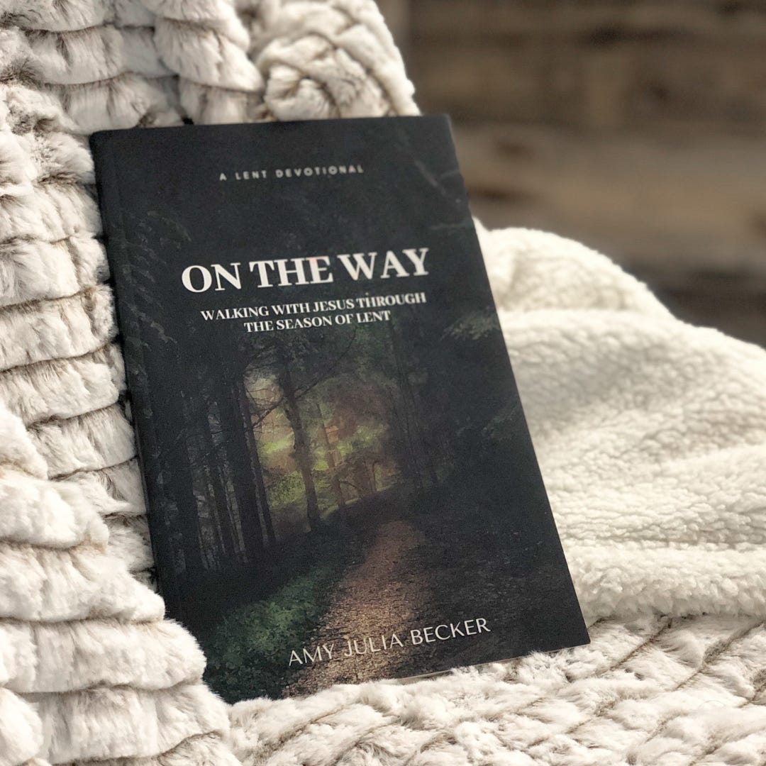 A book titled On the Way: Walking with Jesus Through the Season of Lent by Amy Julia Becker rests on a soft, textured blanket. The book cover features an image of a forest path illuminated by warm, golden light filtering through the trees.