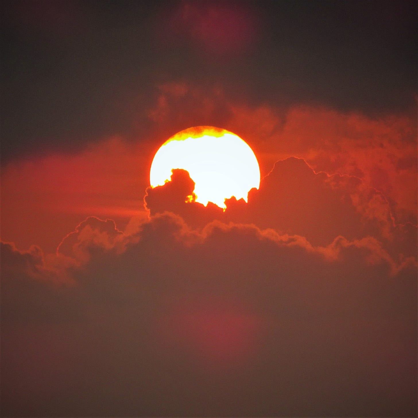 A glowing orange sun sets behind multiple layers of cumulus clouds.