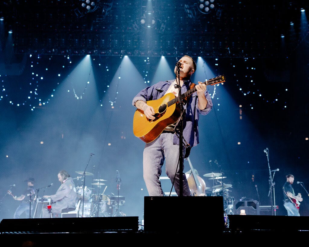 A man in a blue shirt with the sleeves rolled up plays an acoustic guitar and howls into a microphone on a big stage, with a fiddler, guitarist, lap steel player, drummer, bassist and acoustic guitarist visible behind him.