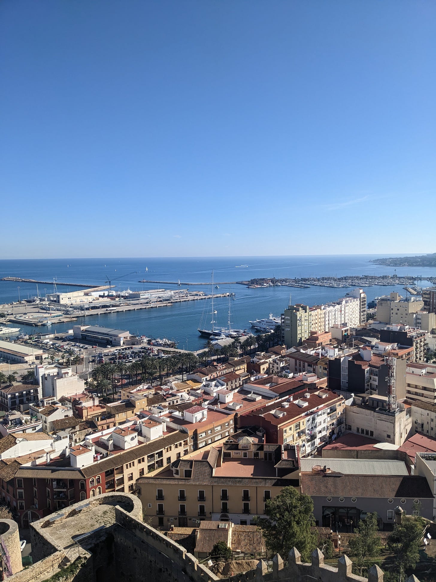 a photo of Dénia and its port taken from above. Blue sky, blue water, boats and buildings.