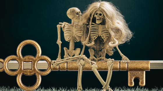 Two skeletons perch on a bench made of wood and a huge key, one with long, flowing doll hair, the other without. They look cozy as they grin together