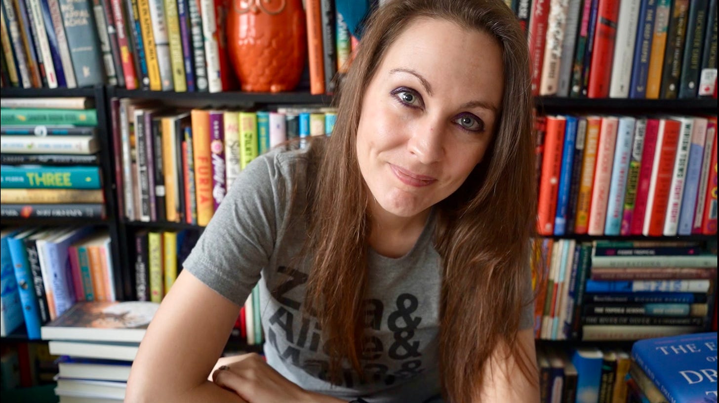 a photo of Kendra, a white woman with brunette hair. She’s posing with her arms crossed. A row of bookshelves can be seen behind her.