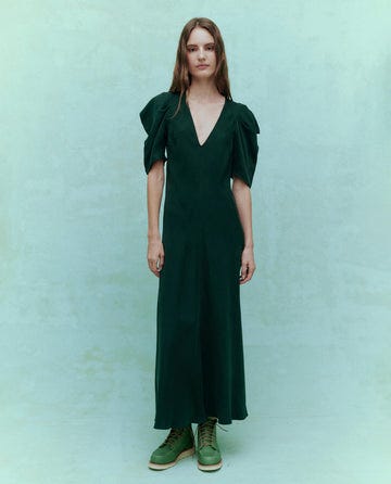 A person stands against a pale green background wearing The Century Dress in Winter Pine by THE GREAT. HOL 24, featuring puffed sleeves and paired with coordinating green boots.