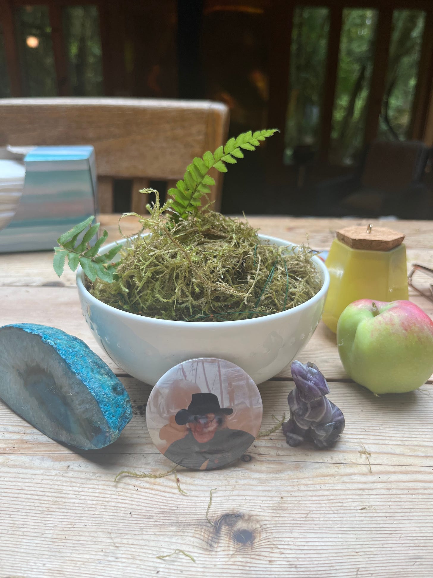 A bowl of moss sits on a table surrounded by small items. Propped up against the bowl of moss is a button pin with a photo of Jack Hirschman.