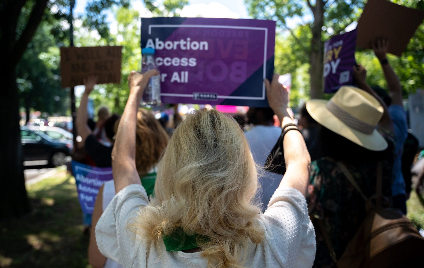 A blond white woman is holding up a sign that says "Abortion Access for All" and marching with other sign holders.